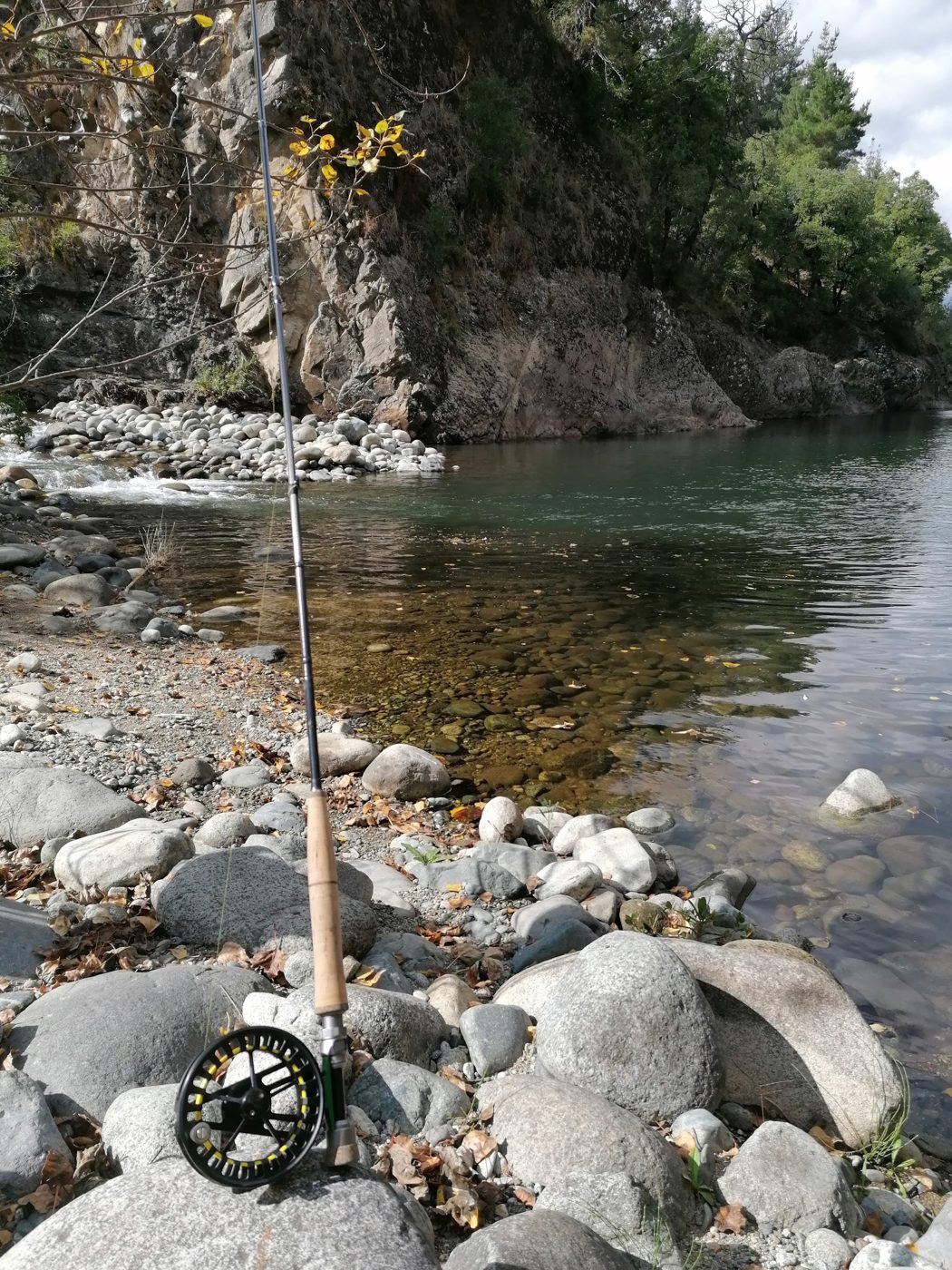 Caña de pesca con mosca y carrete apoyados en las piedras a la orilla de un río de aguas transparentes